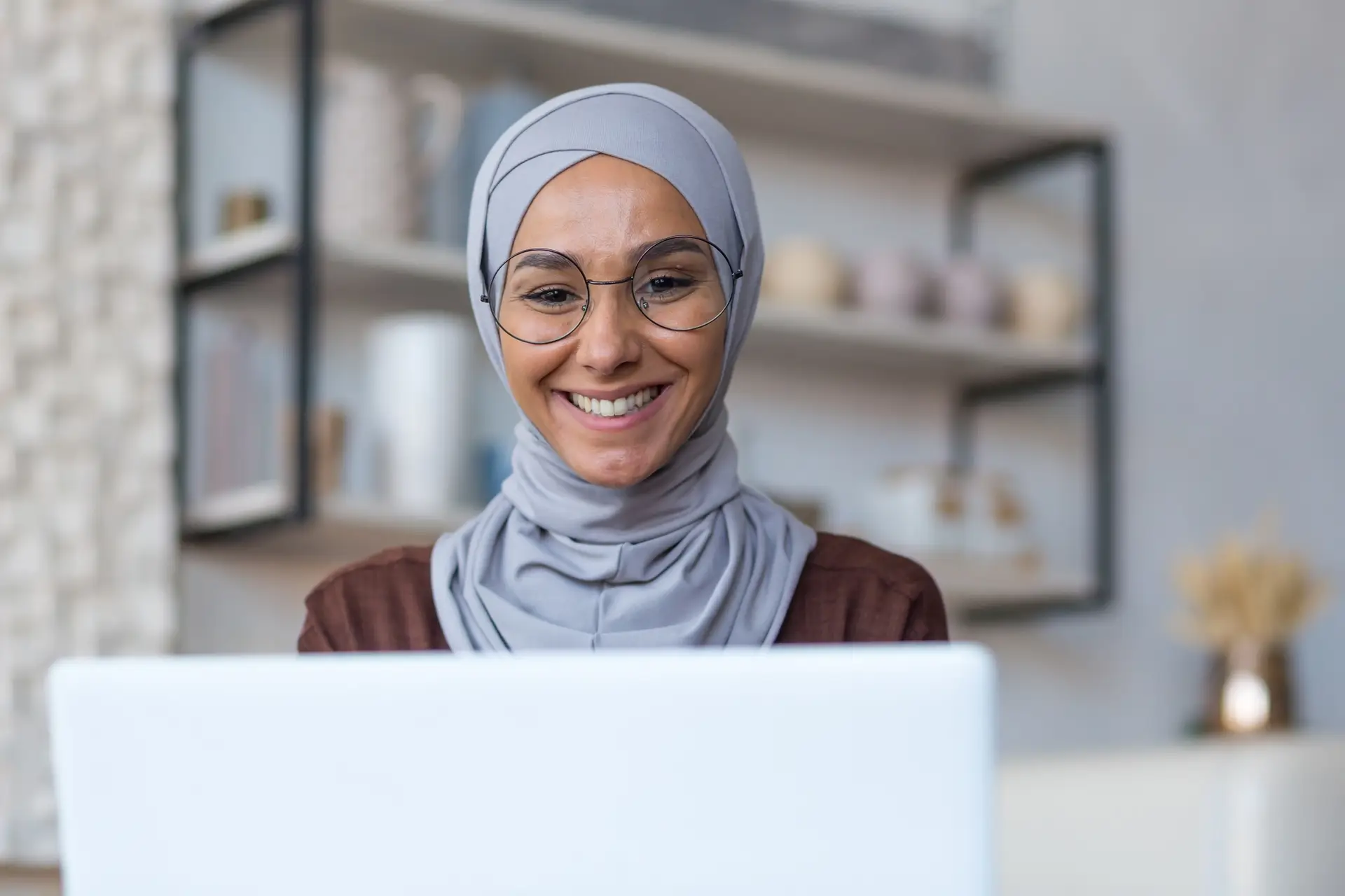 Close-up photo of young beautiful Arab woman in hijab and glasses working at home sitting in kitchen